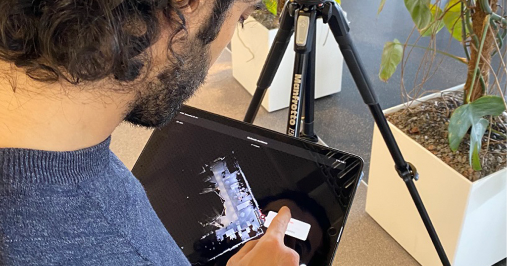 Mann prüft auf einem Tablet den 3D-Scan eines Grundrisses, daneben ein Kamerastativ